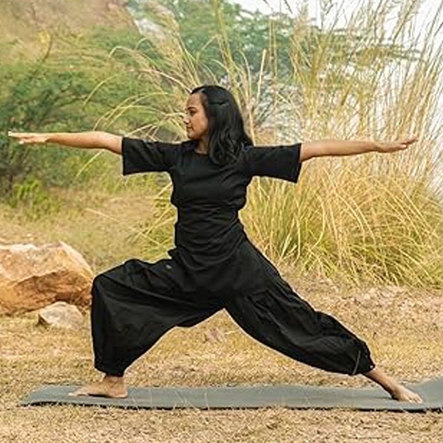 Person in black outfit practicing yoga outdoors with greenery in the background