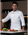 Chef in a white uniform standing in a kitchen with vegetables on a cutting board.