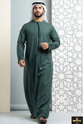 Man wearing a green traditional outfit standing against a white wall with geometric patterns.