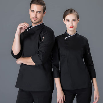 A man and a woman modeling matching black chef jackets with silver-gray trim and silver snap closures, posed against a solid gray studio background.