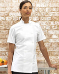 A woman wearing a white short-sleeve double-breasted chef jacket stands in front of a light brick wall, with a table holding fruit visible at the lower left.
